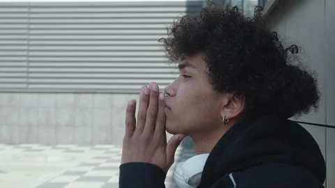 A young afro American guy in a black hoodie sitting near a concrete wall Praying Stock Footage 154355630