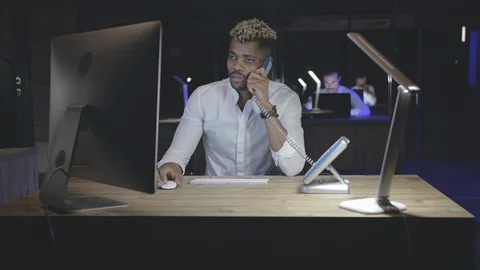 Young afro office worker having phone call and using computer in dark office Stock Footage 125684928