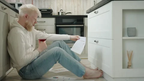 Young albino man, drinking red wine, sitting on floor, holding sheet of paper Stock Footage 244850452