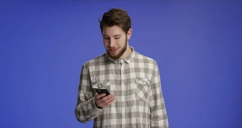 Young and handsome man typing on his phone and smiling in front of dark blue  Stock Footage 99415705