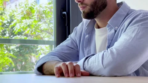 Young anxious man with a beard sitting at a table by the window, tapping his Vídeos de archivo 345117605