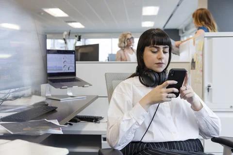 Young apprentice with headphones looking at phone at desk Stock Photos