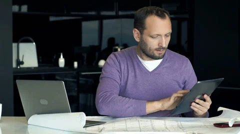 Young architect comparing tablet and blueprints by table in the kitchen Stock Footage 62623191