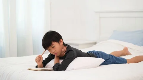Young Asian boy lay on a bed and get an idea to solve homework Stock Footage 129553693