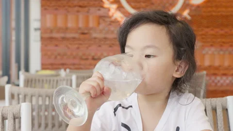 Young Asian child drinks refreshing iced water from a large glass while sitting Stock Footage 338406640
