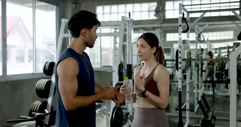 Young Asian couple talks in modern gym while sharing water bottle, smiling 動画素材 342585961