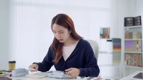 Young Asian designer sketching notes while examining fabric swatch at brigh.. Stock Footage 345149046