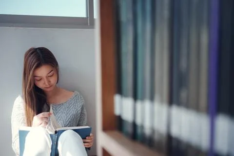 Young asian female college student sitting on floor in library, reading book. Stock Photos