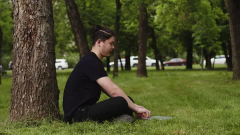 Young asian guy sitting under the tree outdoors in the city park on summer Stock Footage 246362879