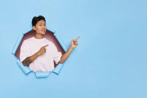 Young Asian guy standing inside the hole of blue background pointing fingers Stock Photos