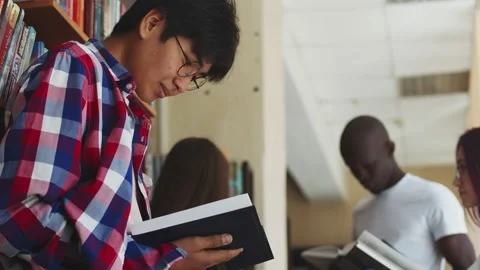 Young asian guy student reading a book in a library. Black guy and caucasians Stock Footage 221411593