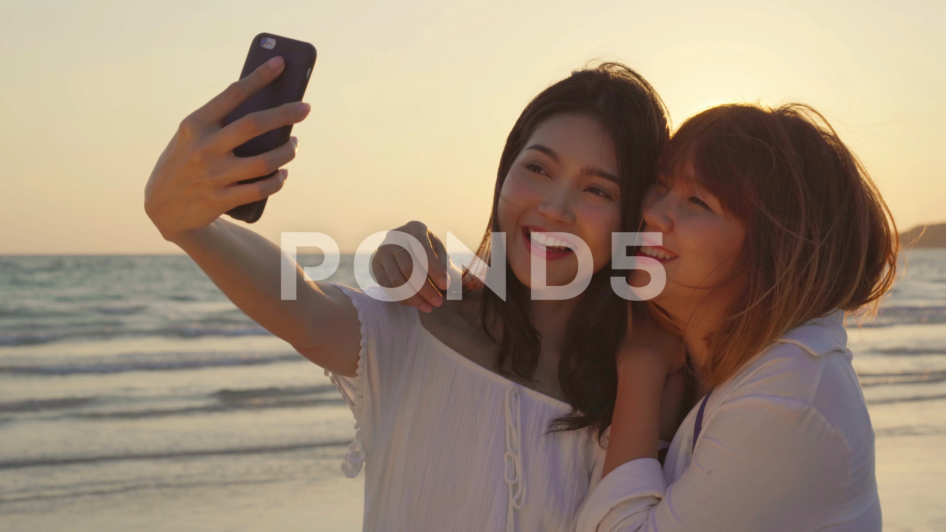 Young Asian lesbian couple using smartphone taking selfie near beach.