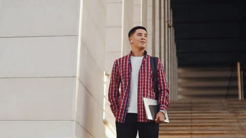 Young asian man with a backpack and laptop stands outdoors on the stairs and Stock Footage 253719656
