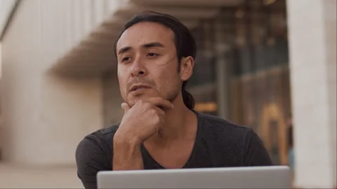 Young asian man close up working on a laptop outdoors with a thoughtful and Stock Footage 253059014