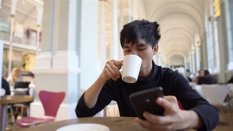 Young Asian Man Drinks Hot Beverage in Cafe or Restaurant And Texting on Phone Stock Footage 117969953