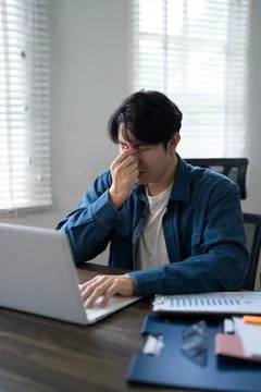 Young Asian man experiencing work stress rubbing eyes while working on lapt.. 写真素材