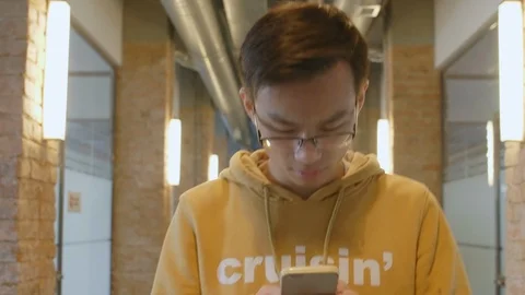Young Asian Man Going through Hallway and Typing on Smartphone Stock Footage 126135313