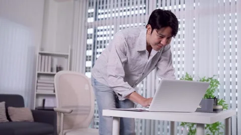 Young Asian man leaning over desk typing on laptop in bright modern home offi Stock Footage 345156327