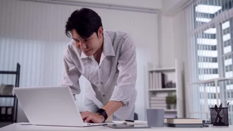 Young Asian man leaning over desk typing on laptop in bright modern home of.. Stock Footage 345156635