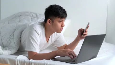 Young Asian man lying in the bed and using smartphone and laptop Stock Footage 156639316