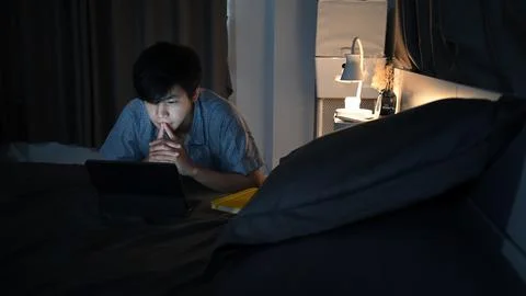 Young asian man lying on his bed and surfing inter.net with computer laptop. Stock Photos