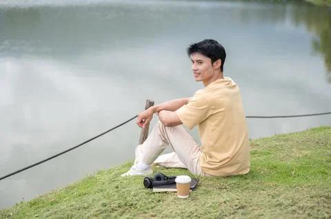 Young Asian man relaxing while sitting on grass with coffee beside a scenic.. Stock Photos