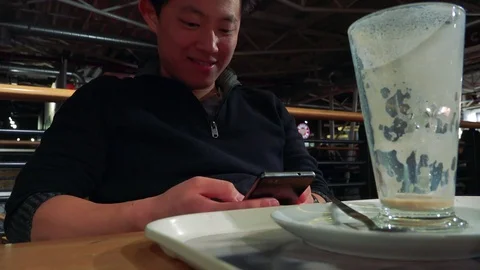 A young Asian man sits at a table in a cafe and works on a smartphone, a tall 스톡 동영상 77299415