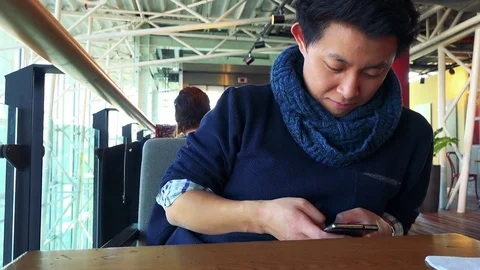 A young Asian man sits at a table in a cafe and works on a smartphone with a Stock-Footage 77300676