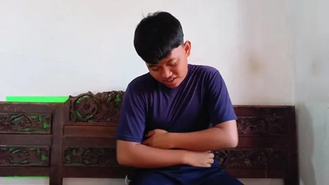 A young Asian man is sitting down, clutching his stomach and expressing pain. Video stock 321938900