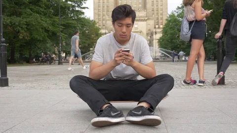 Young Asian Man sitting on his skateboard in a park Stock Footage