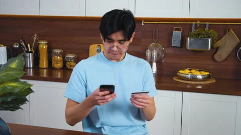 Young asian man sitting at modern kitchen, involved using smart phone for online Stock Footage 162569982