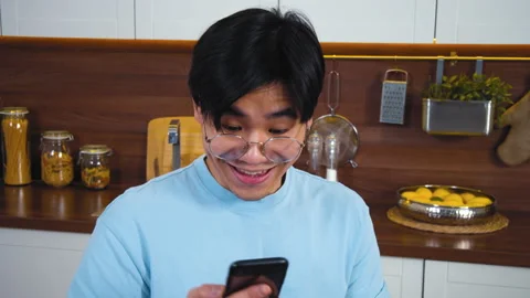 Young asian man sitting at modern kitchen, involved using smart phone for online Stock Footage 162571925