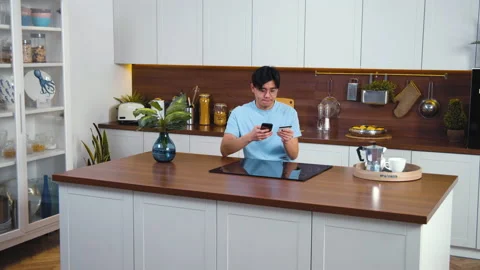 Young asian man sitting at modern kitchen, involved using smart phone for online Stock Footage 165783204