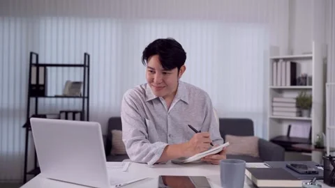 Young Asian man smiling and writing notes at laptop desk in bright hybrid w.. Stock Footage 345156584