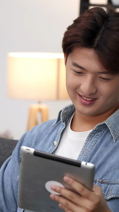 Young Asian Man Smiling While Using Tablet Computer on Living Room Sofa 動画素材 329283767