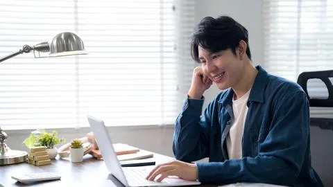 Young Asian man smiling while working on laptop at bright modern home offic.. 写真素材