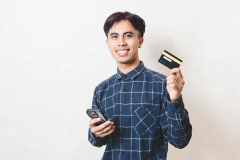 Young Asian man smiling while holding a smartphone and credit card, represe.. Stock Photos