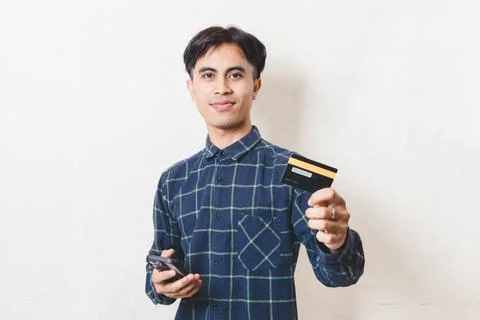 Young Asian man smiling while holding a smartphone and credit card, represe.. Stock Photos