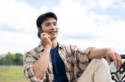 Young Asian man smiling while talking on smartphone on bench beside a mount.. Stock Photos
