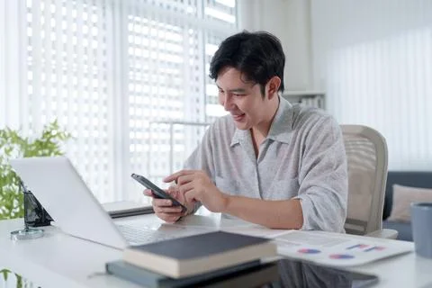 Young Asian man smiling while using smartphone and laptop working from brig.. Stock Photos
