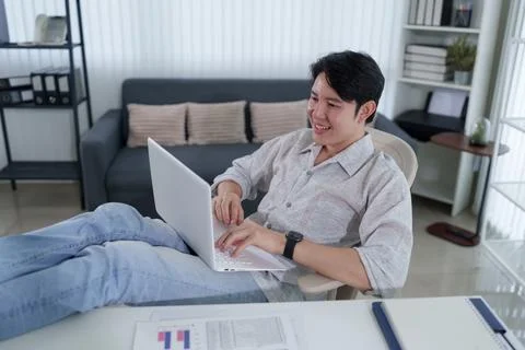 Young Asian man smiling while working on laptop in modern living room durin.. Stock Photos