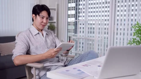 Young Asian man taking notes on tablet during video call at home office des.. Stock Footage 345156338