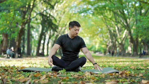 A young asian man in a tracksuit is doing yoga on a mat in a city park using Stock Footage 232988302
