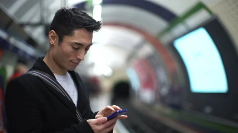 Young asian man types on his phone at a subway station Stock Footage