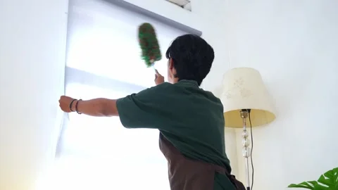 Young asian Man uses feather duster to clean white roller blinds in home Stock Footage 327446076