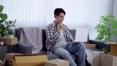 Young Asian man using laptop while sitting on couch surrounded by moving bo.. Stock Footage 341546908