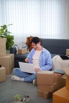 Young Asian man using laptop while sitting on floor surrounded by cardboard.. Fotos de archivo