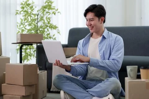 Young Asian man using laptop while sitting among packed cardboard boxes in .. Stock Photos