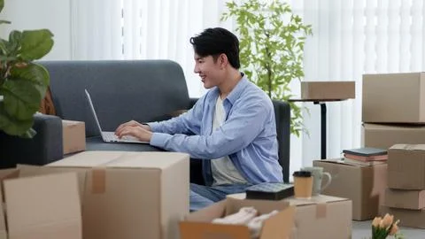 Young Asian man using laptop to manage home relocation surrounded by cardbo.. Stock Photos