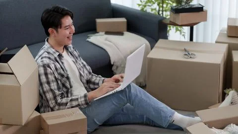 Young Asian man using laptop computer while sitting on floor surrounded by .. Stock Photos